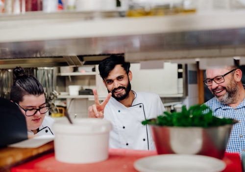 Drei Männer stehen in der Küche, der mittlere zeigt ein Peace-Zeichen